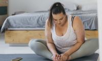 En større ung kvinde sidder på sin yogamåtte og kigger ned på sine samlede fødder.